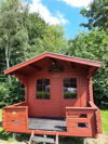 Rote Holzhütte mit kleiner Terrasse vorne, umgeben von grünen Bäumen und Sommerhimmel.